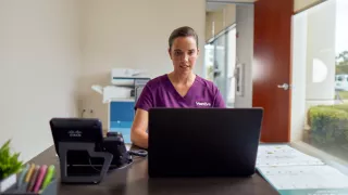A healthcare professional in scrubs sits at her desk with her laptop open