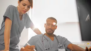Two healthcare professionals in scrubs make notes as they watch an online learning course at their desk