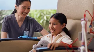 A child is in a chair connected to the AK 98 Dialysis Machine, with a nurse at the arm rest, looking at her and smiling.