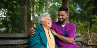 Healthcare professional and a patent sit on a bench in an outdoor space with trees in the background
