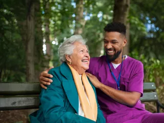 Healthcare professional and a patent sit on a bench in an outdoor space with trees in the background