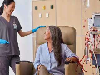 A woman in a chair is connected to the AK 98 Dialysis Machine, and conversing with a nurse standing beside her
