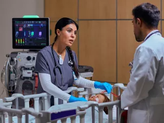 Healthcare professional leans over a bassinet and has a discussion with a colleague. ThePrisMax system stands behind her.