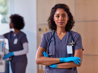 A healthcare professional in scrubs and blue gloves looks straight on with a smile and her arms crossed.